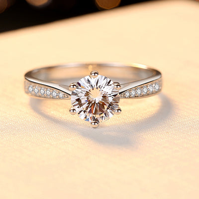 Shimmering silver ring plated with white gold, featuring a 1 CT round cut moissanite diamond held securely by six prongs, surrounded by delicate diamond accents in a channel setting.